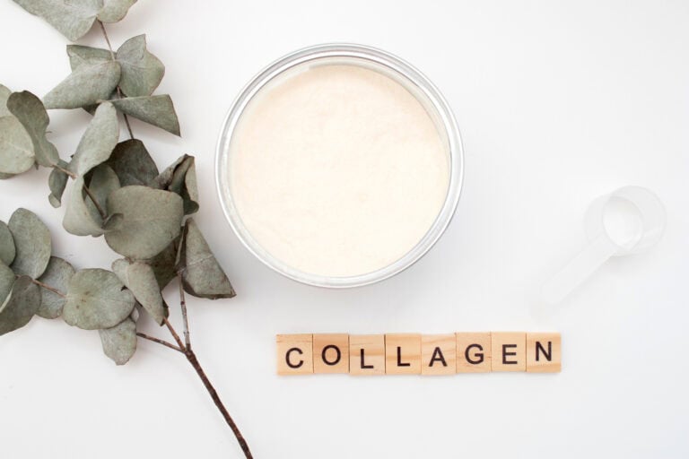 Open container of collagen powder with a scoop and the word "collagen" spelled out in letter tiles, accompanied by eucalyptus leaves on a white background.