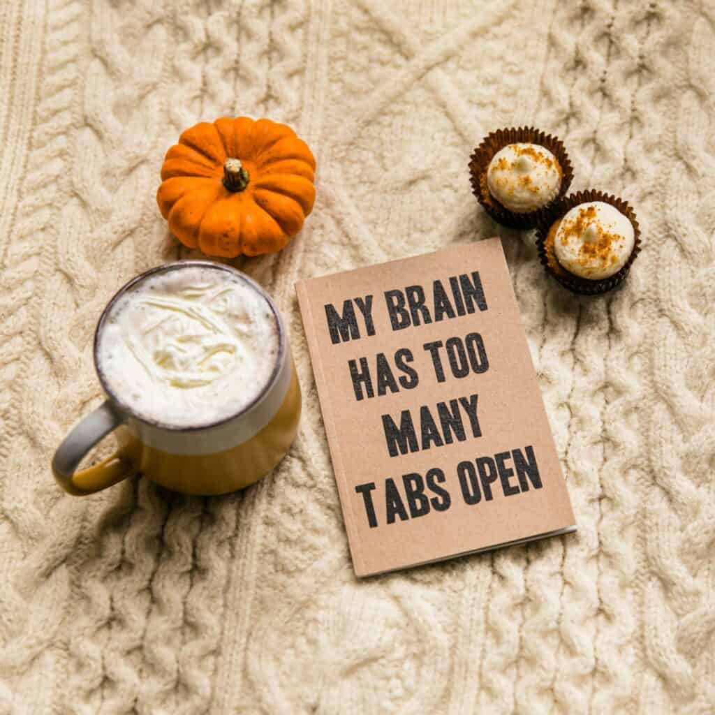 A cozy autumnal setting with a latte, a mini pumpkin, two bite-sized desserts, and a humorous note stating, "my brain has too many tabs open.