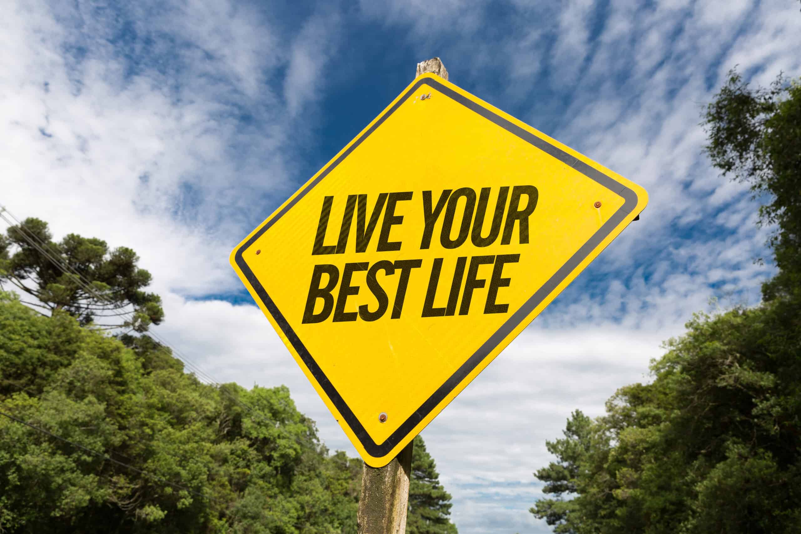 A yellow road sign with the phrase "live your best life" against a backdrop of blue sky and trees.