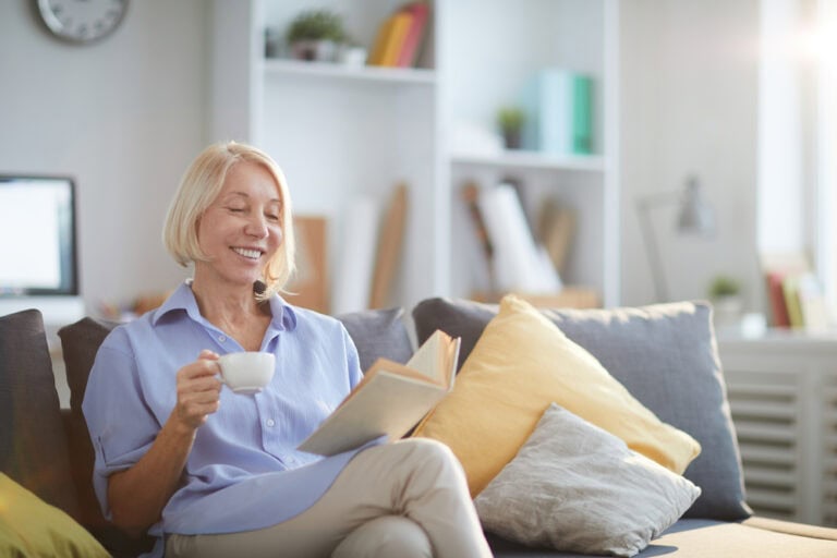 Woman smiling while reading a book and holding a cup of coffee on a cozy sofa.