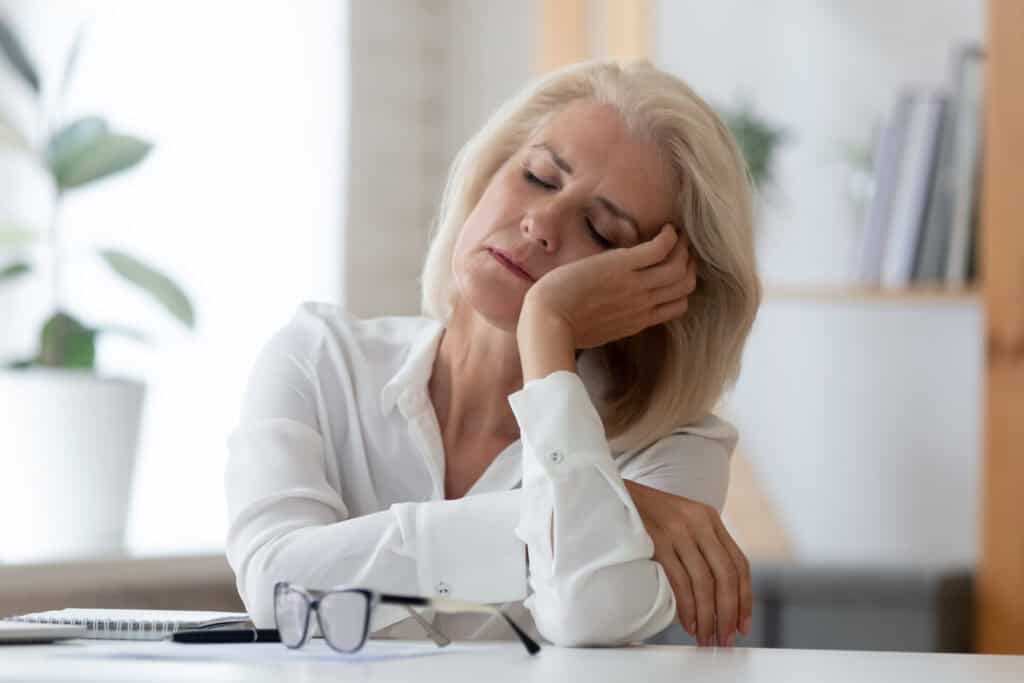 A tired middle-aged woman resting her head on her hand at a desk, appearing exhausted or stressed.