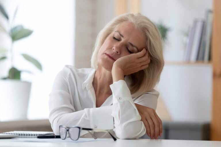 A tired middle-aged woman resting her head on her hand at a desk, appearing exhausted or stressed.