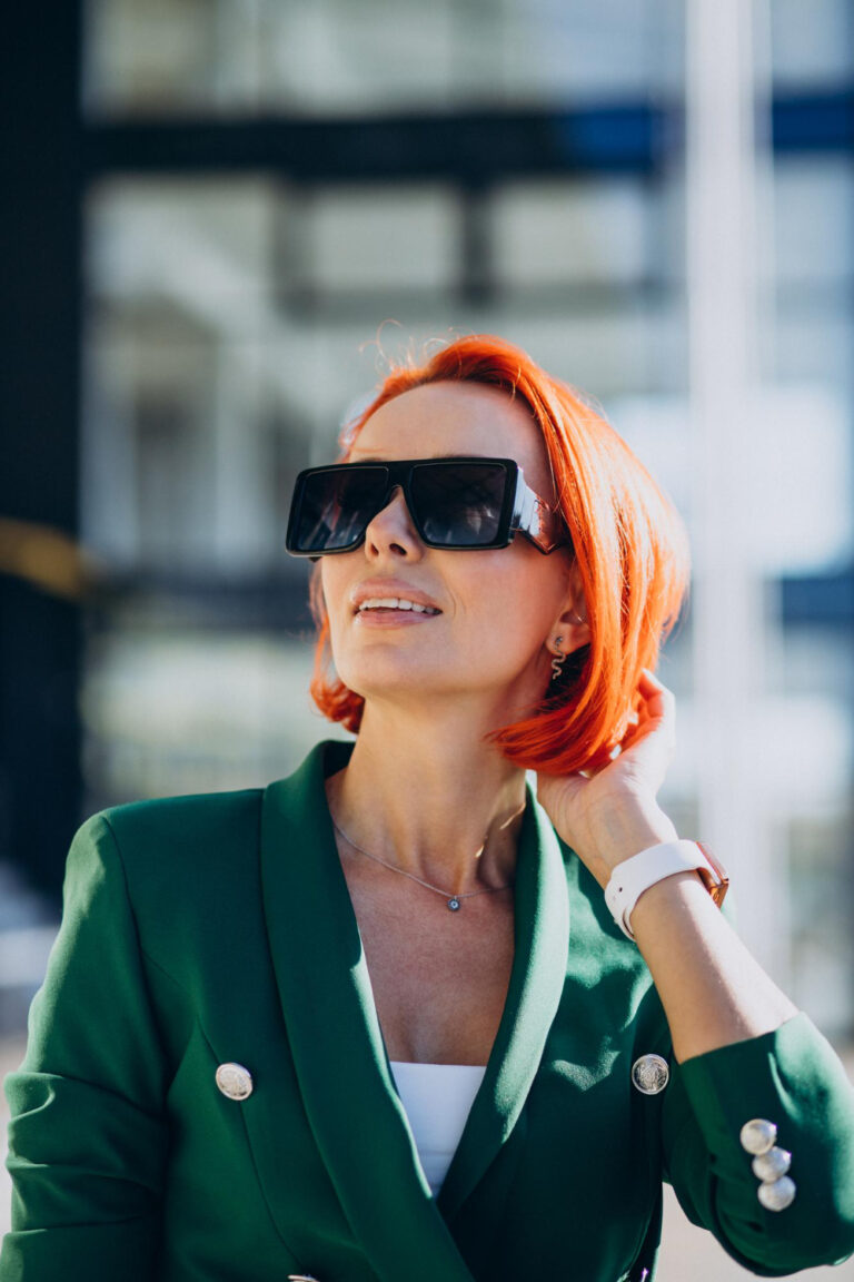 Woman with bright red hair wearing sunglasses and a green blazer looks up, thoughtful, in a sunlit urban setting.