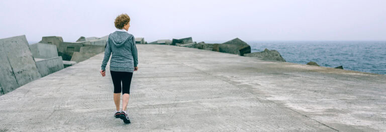 A person in a gray hoodie and black leggings walks alone on a concrete pier with the ocean and large rocks in the background.