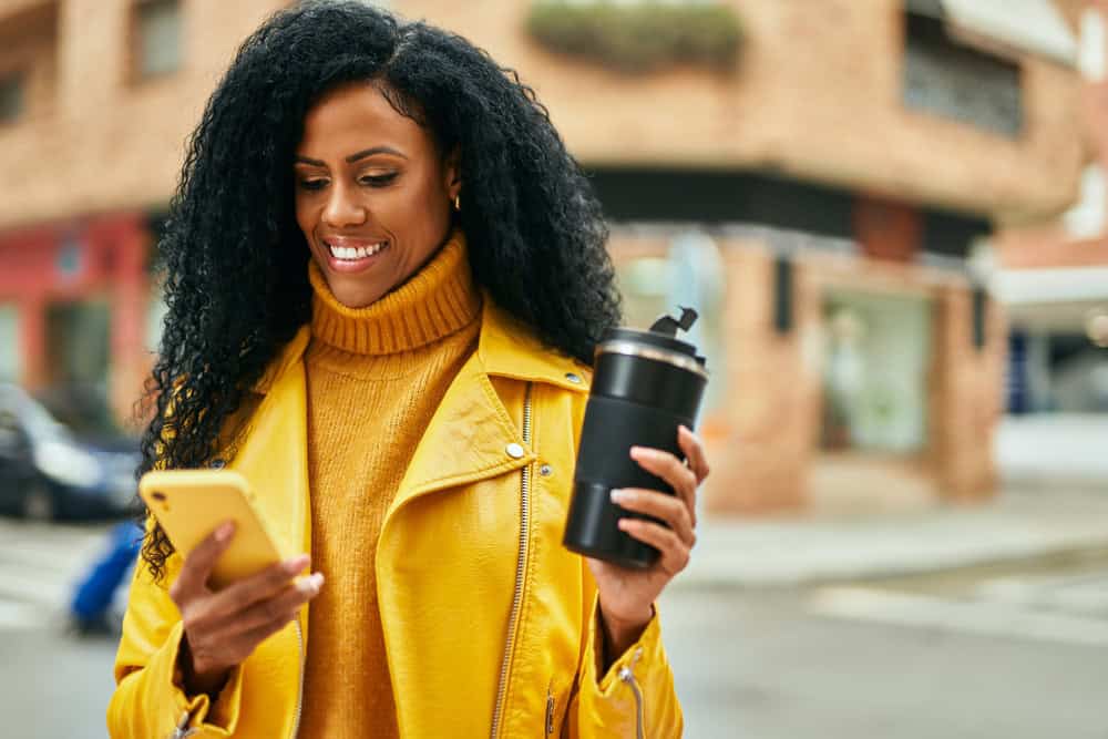 Woman wearing a yellow jacket holds a travel mug in one hand and a smartphone in the other, smiling as she looks at her phone on a city street.