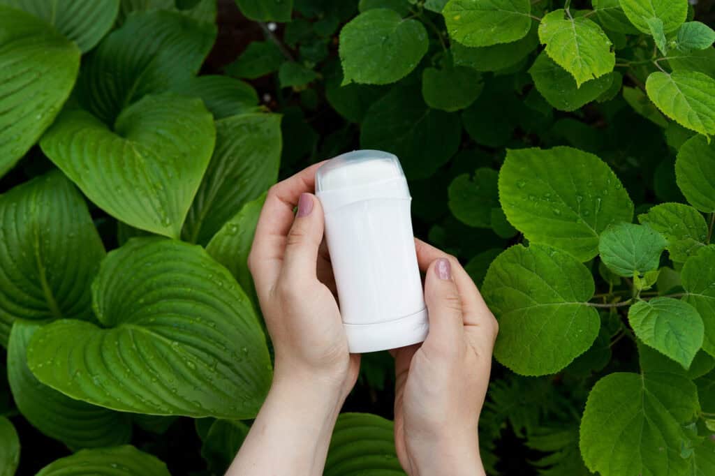 Hands holding a white deodorant stick in front of green leaves.